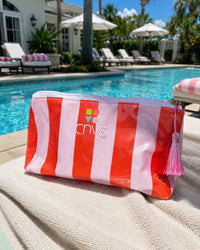 A pink and red striped cosmetic pouch with a pink tassel zipper pull, featuring the 'cnvs' logo, resting on a beige towel in front of a swimming pool.