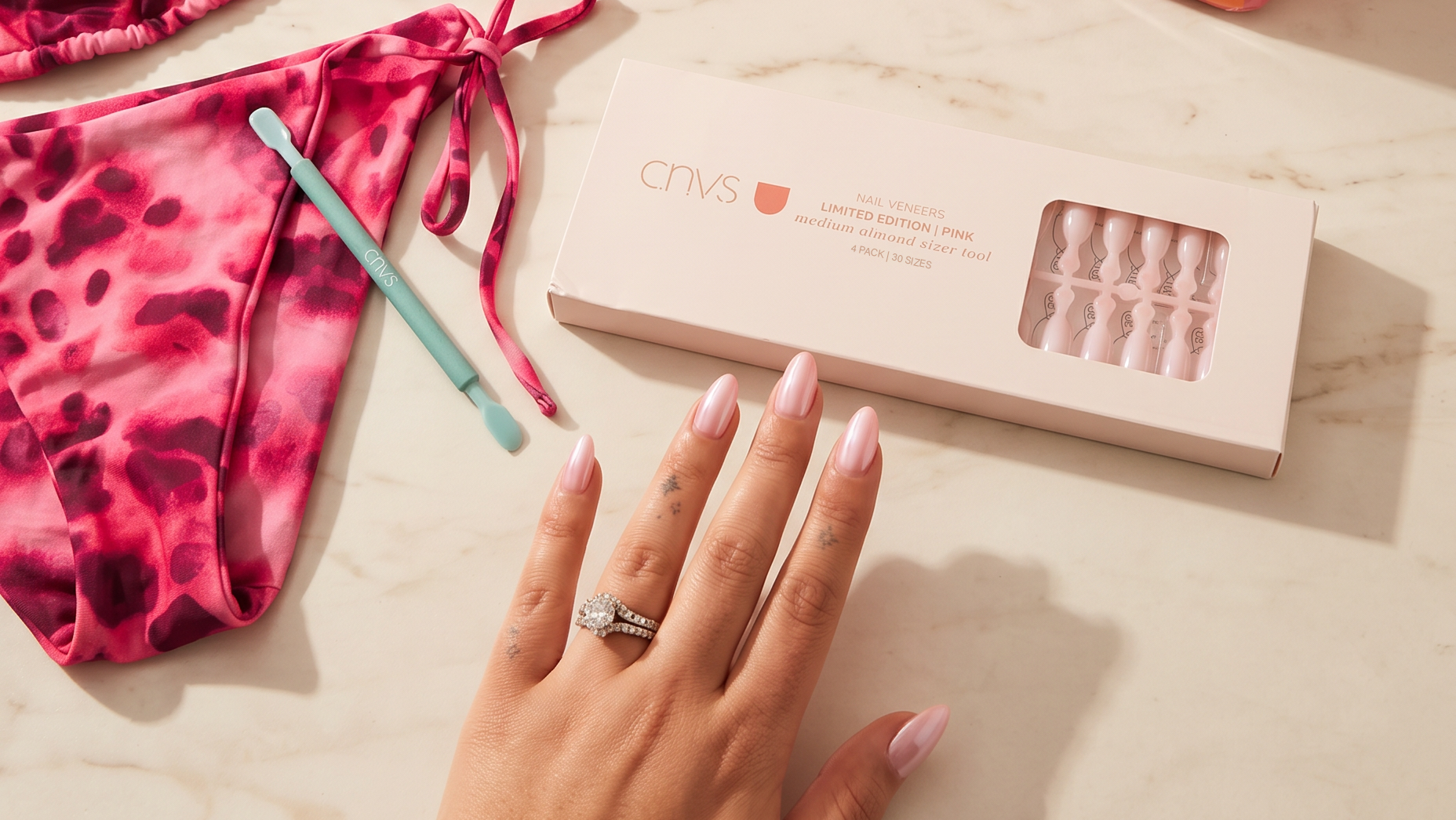 A styled flat-lay featuring a pink and magenta tie-dye bikini set, a large orange and pink striped CNVS tote bag, a box of CNVS press-on nails, sunglasses, a drink, and a hand resting on a light marble surface.